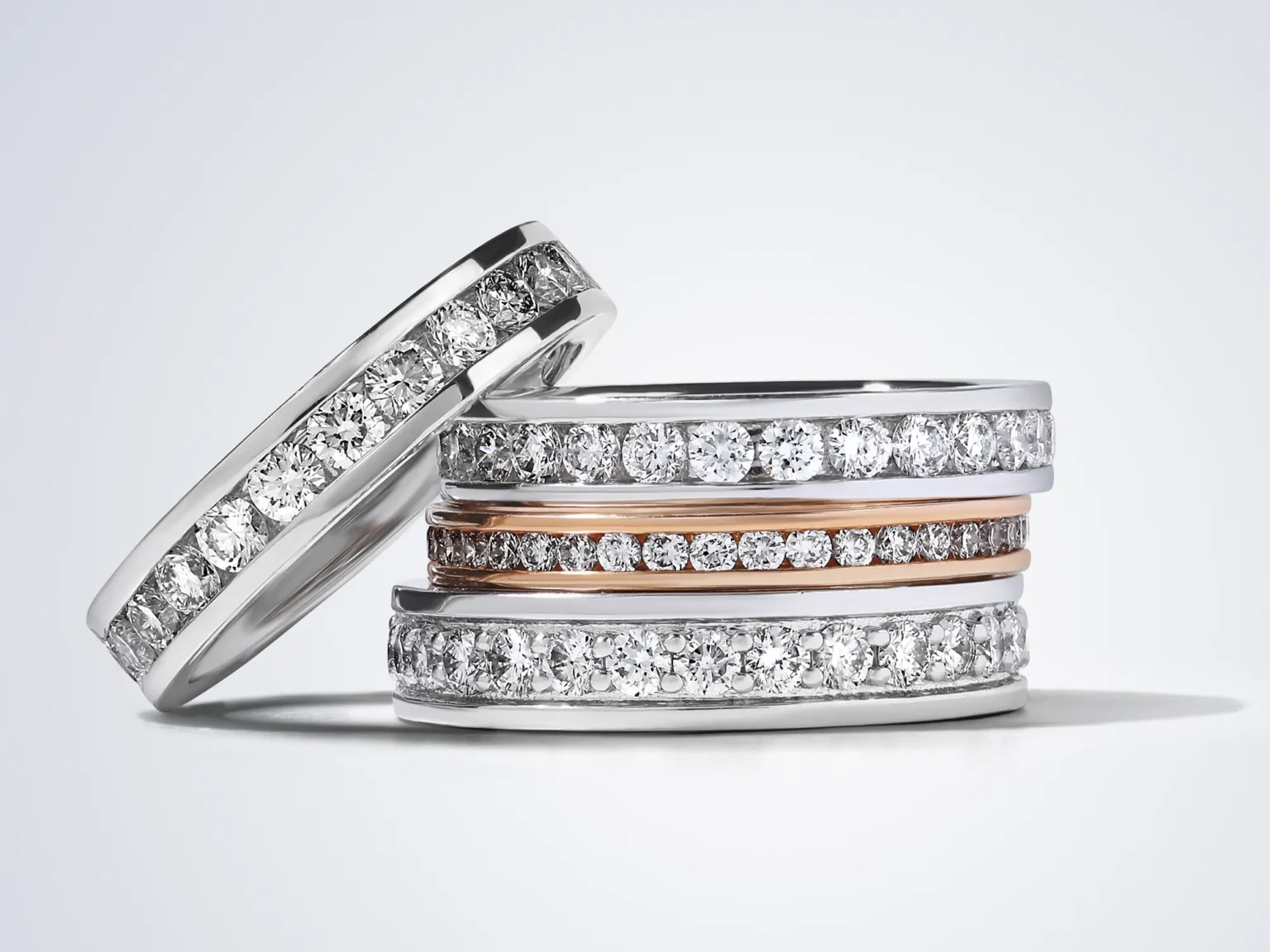 A stack of three white and rose gold diamond eternity bands on a soft grey background.