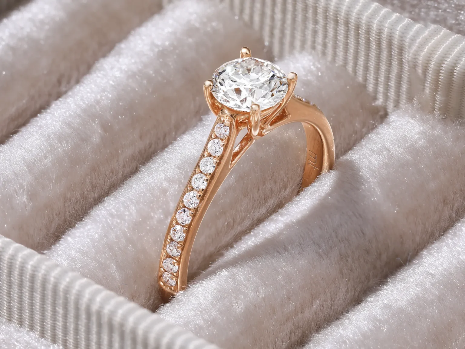 Close-up of a rose gold diamond engagement ring with a pav&eacute; band set in a light beige velvet jewellery box.