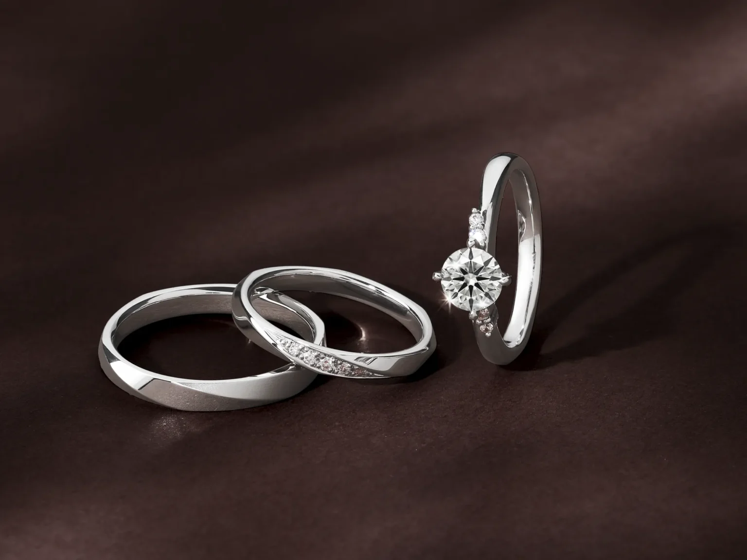 A white gold diamond engagement ring and two wedding bands displayed on a textured dark brown fabric background.