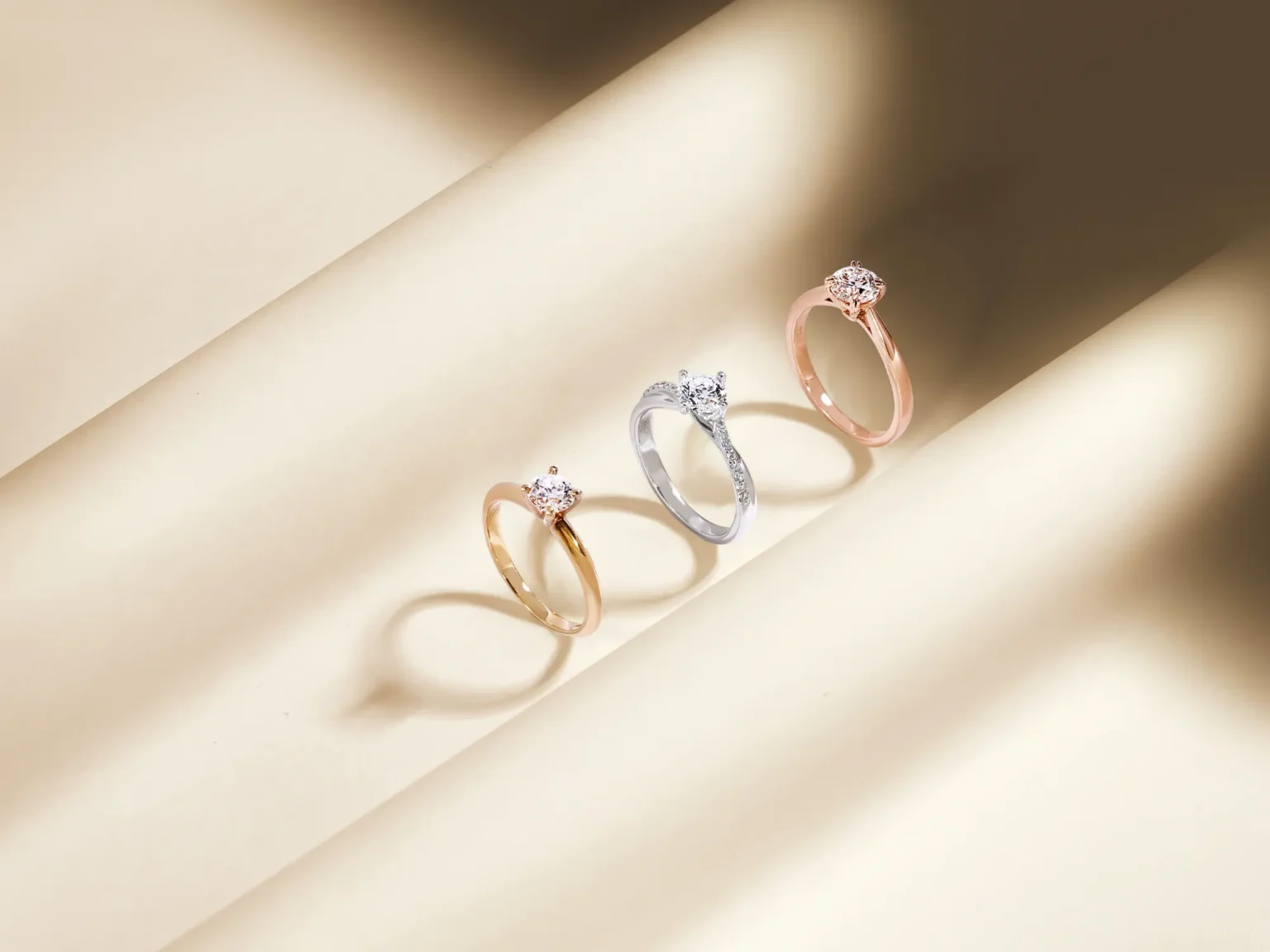 Yellow gold, white gold, and rose gold diamond engagement rings displayed on a wavy, light champagne-coloured surface.