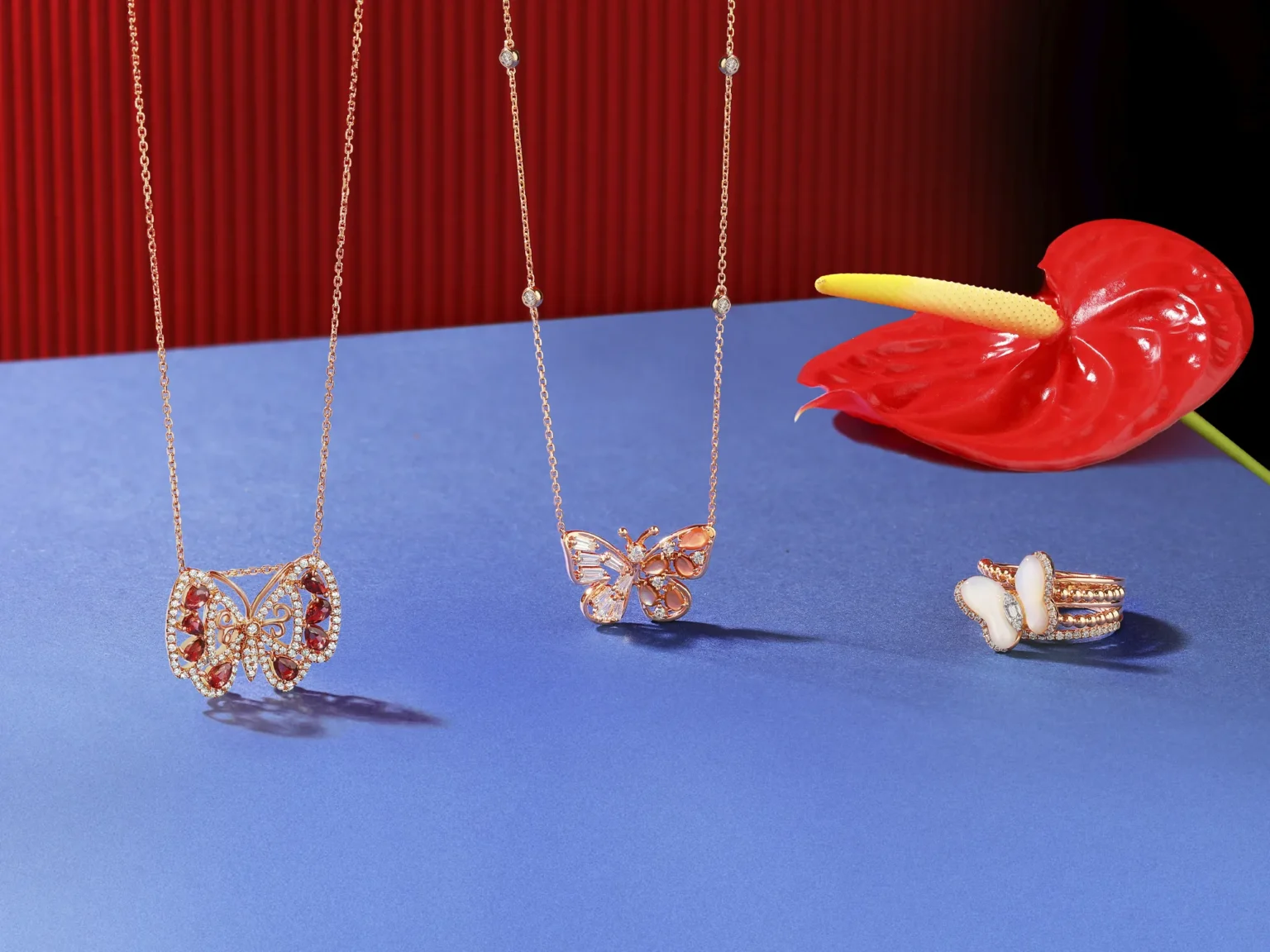 Rose gold butterfly necklaces and a ring with red gemstones and mother-of-pearl styled with a red anthurium on a blue and red background.
