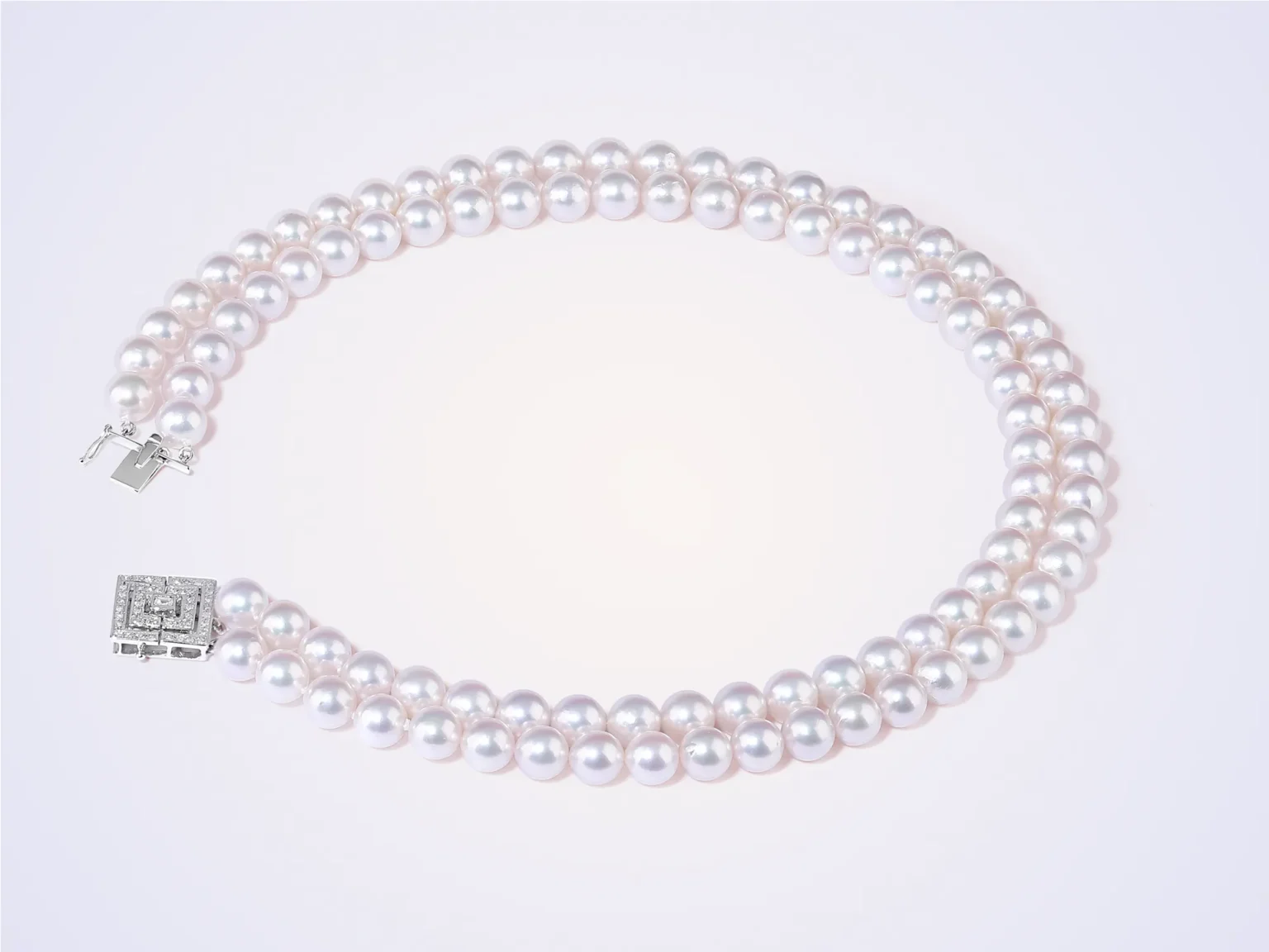 A two-strand white pearl necklace with a square diamond-set clasp on a soft pink background.