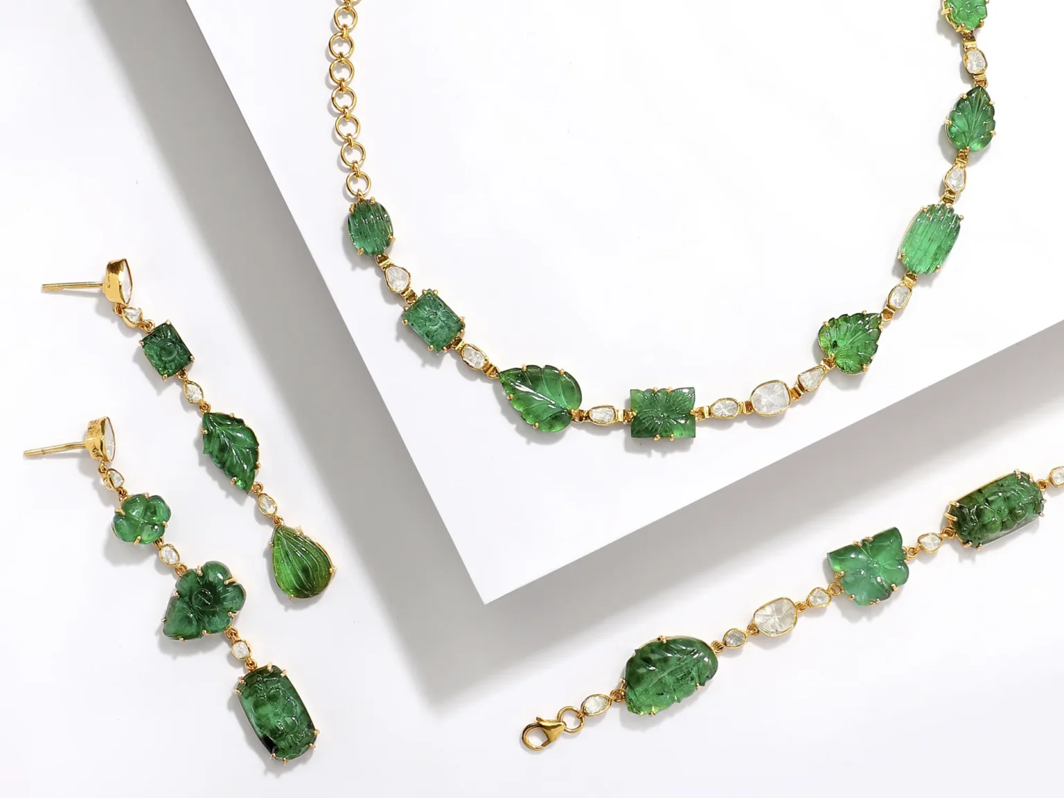 A luxury jewellery set featuring carved emerald leaves and Polki diamonds in yellow gold, including a necklace, bracelet, and earrings on a white background.