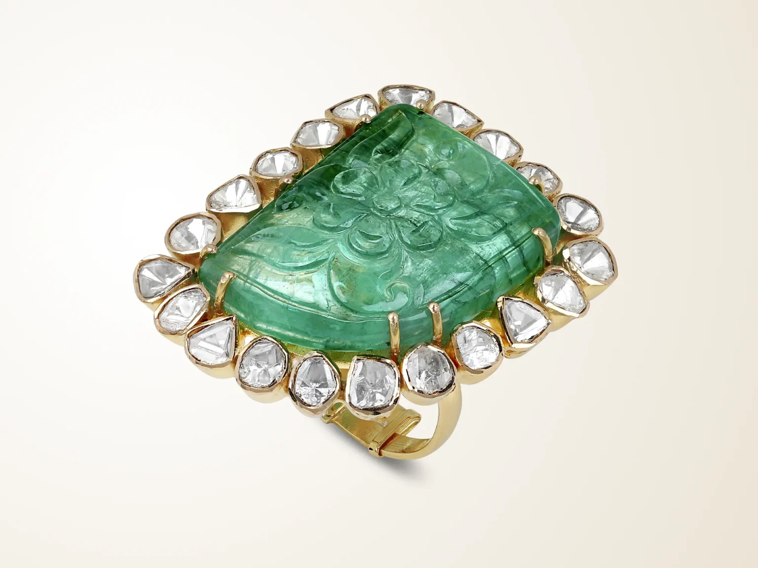 A large 18K gold statement ring featuring a carved floral emerald centerpiece surrounded by a halo of Polki diamonds on a cream background.