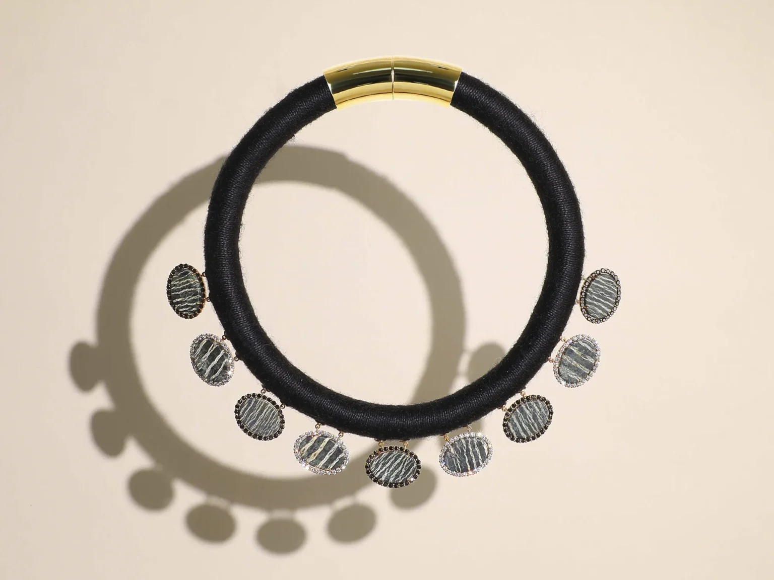 A black silk cord torque necklace with ten oval banded agate and diamond pendants on a beige background with sharp shadows.