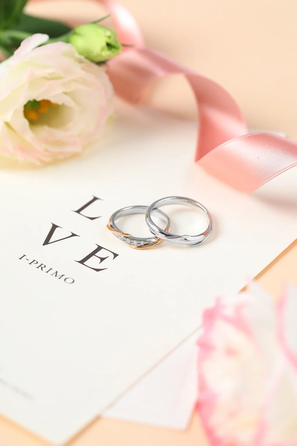 A pair of I-PRIMO platinum and rose gold wedding bands on a "LOVE" card with pink flowers and a silk ribbon.